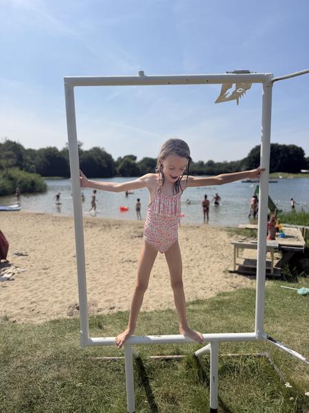 A young girl in a swimsuit joyfully poses within a white frame on a sandy beach, with a lake and lush green trees in the background